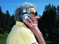 elder­ly woman lis­te­ning to head­phones. exte­rior day park setting